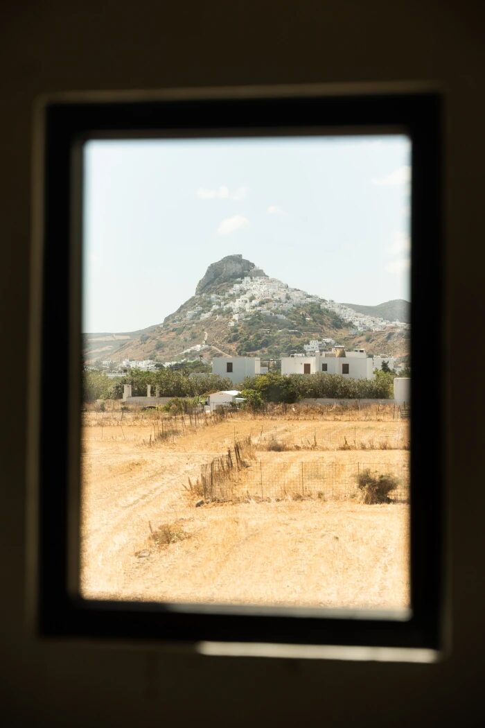 View of Chora village through the bedroom window — whitewashed houses climbing the hill below the Venetian castle, framed by black-steel window